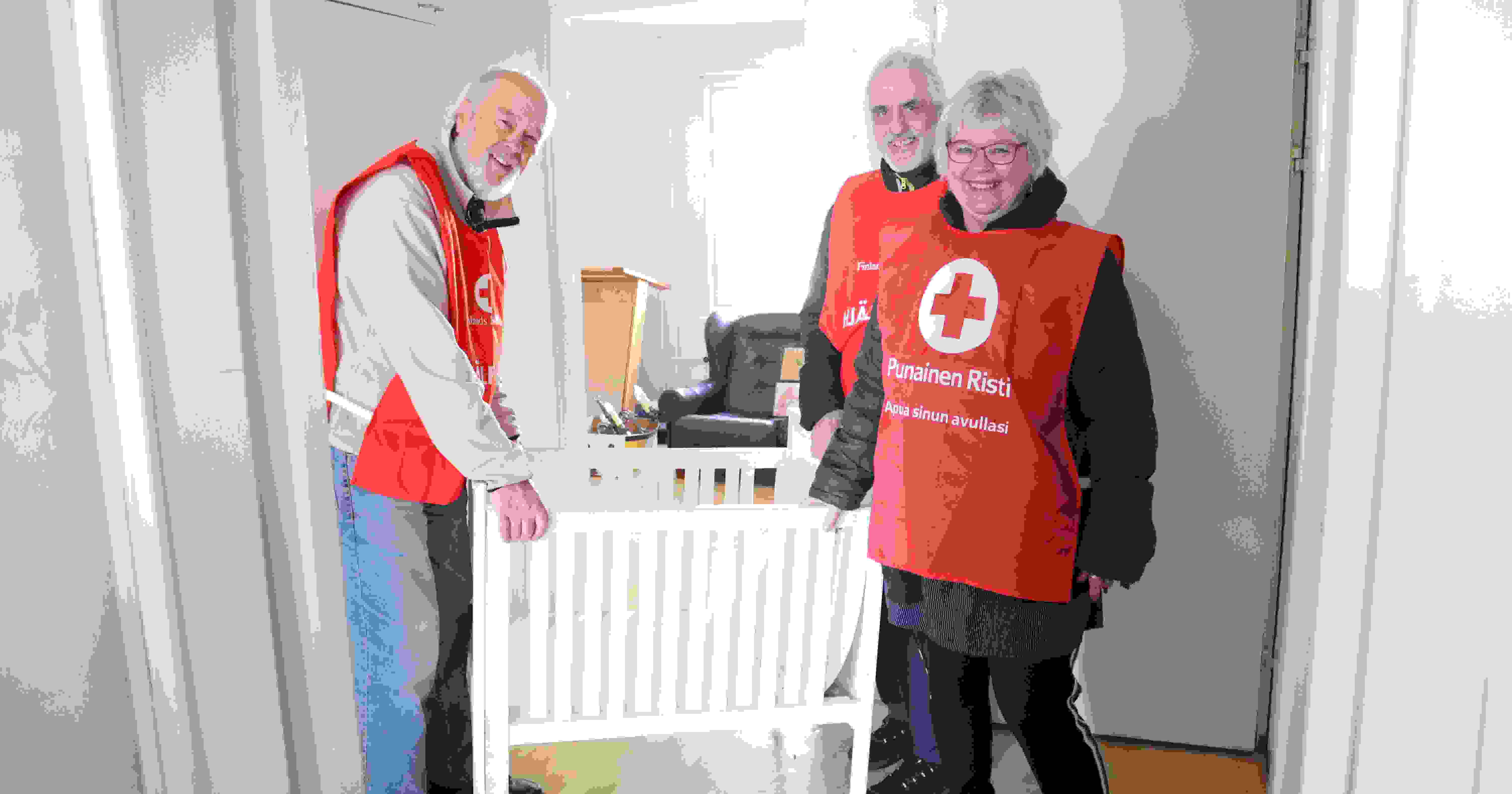 Three smiling people wearing Red Cross volunteer vests stand next to an infant bed in a dwelling that they have furnished for refugees.