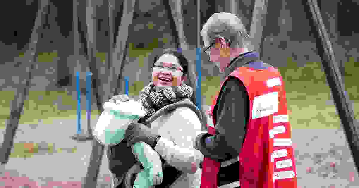 A woman wearing a Red Cross volunteer vest guiding an elderly immigrant woman and child. The elderly woman is hugging the volunteer.