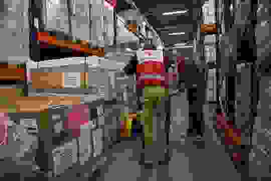 Two Red Cross employees picking up aid supplies in the Logistics Centre, which has wide, tall shelves full of supplies.