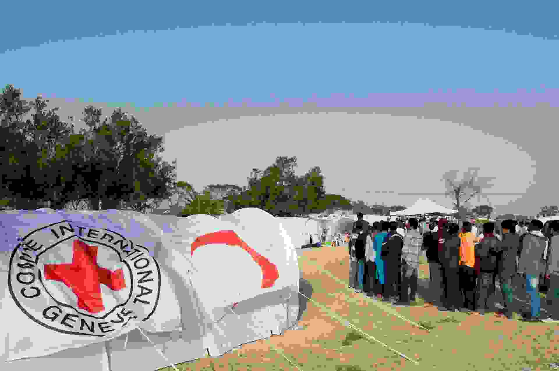 People queueing at a refugee camp among tents equipped with the Red Cross symbols.