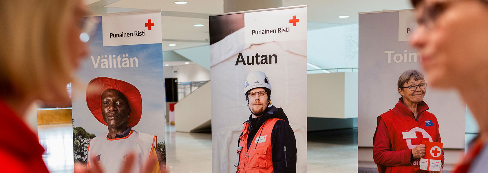 Two people are talking. In the background, there are three roll-ups with Red Cross volunteers.