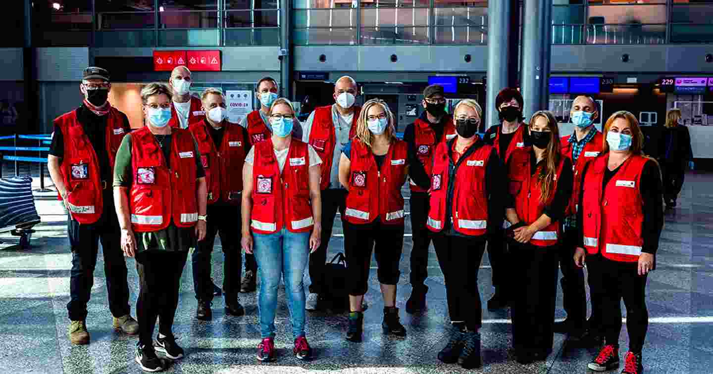 Fourteen aid workers wearing masks and Red Cross gear at Helsinki Airport.