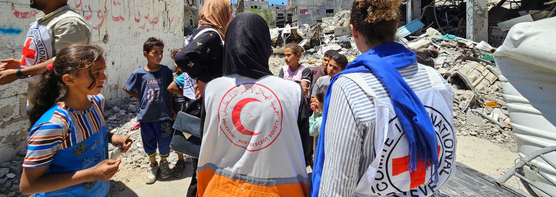 Red Cross and Red Crescent workers talking with children in a destroyed area of Gaza. Collapsed buildings are visible in the background.