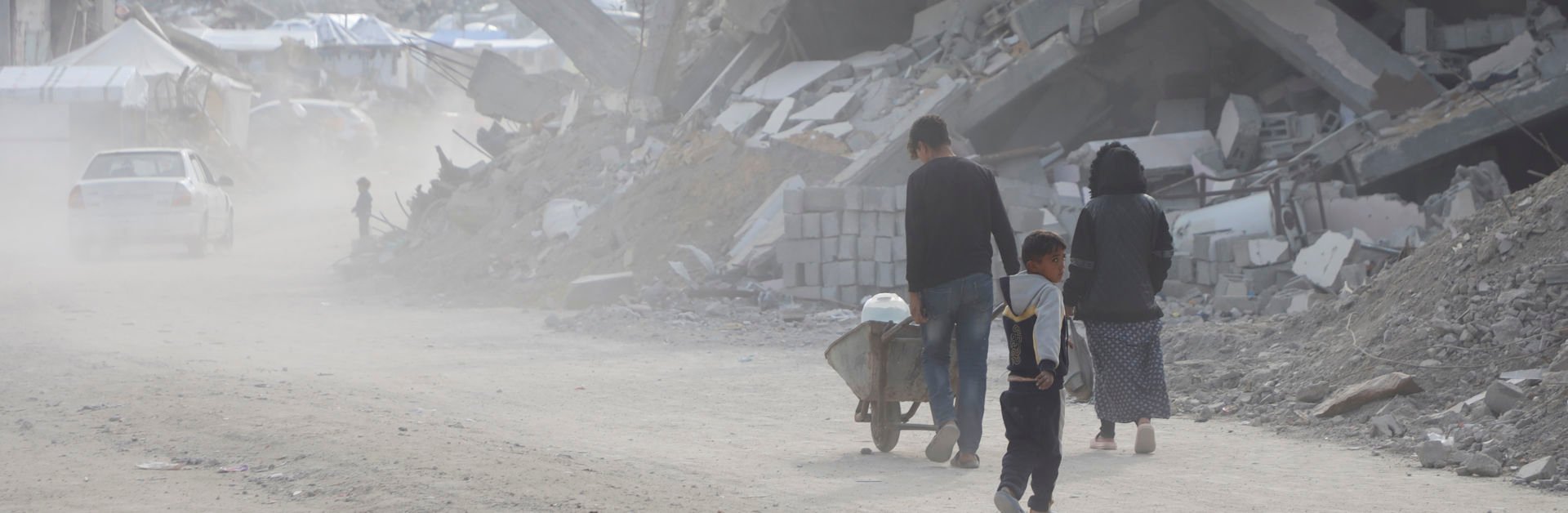A family walks through the rubble in Gaza, one of them pushing a wheelbarrow while a child walks beside, with collapsed buildings in the background.