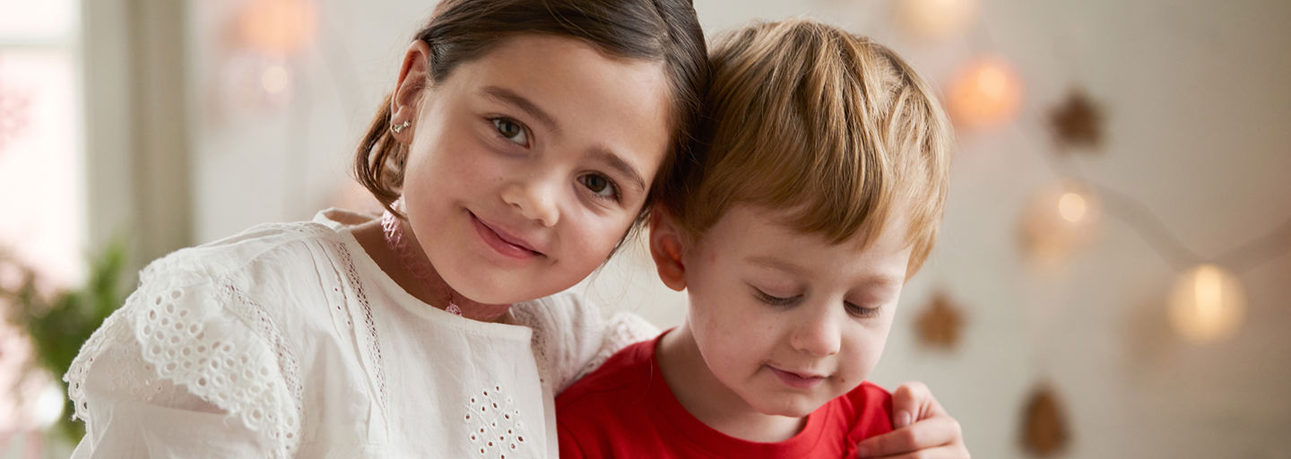 A smiling girl with her hand on her little brother’s shoulder in a room decorated for Christmas.