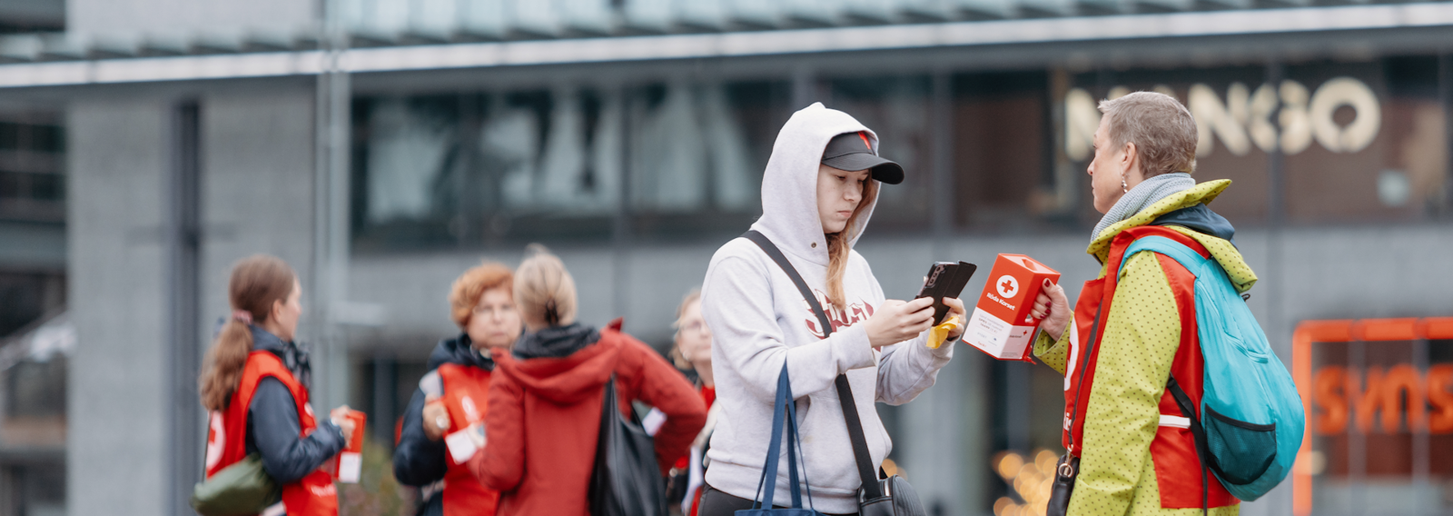 A woman dressed in the Red Cross red vest standing in an autumn view with a collection box in her hand.