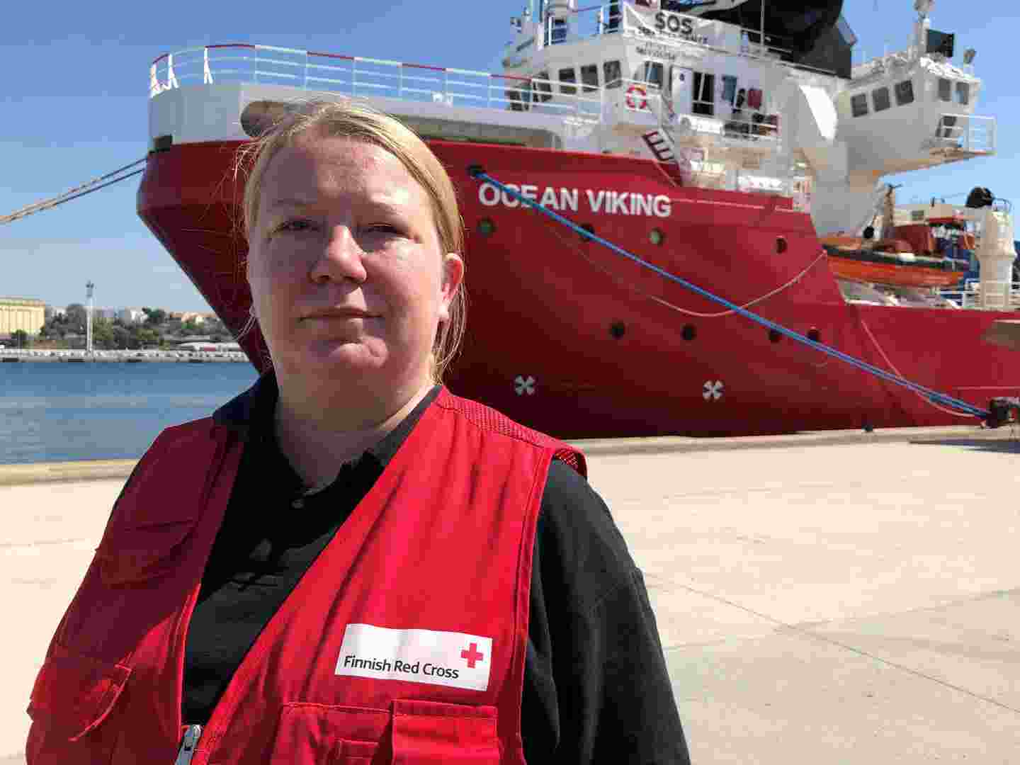 Aid worker wearing Red Cross work clothing standing in front of a ship in a harbour area.
