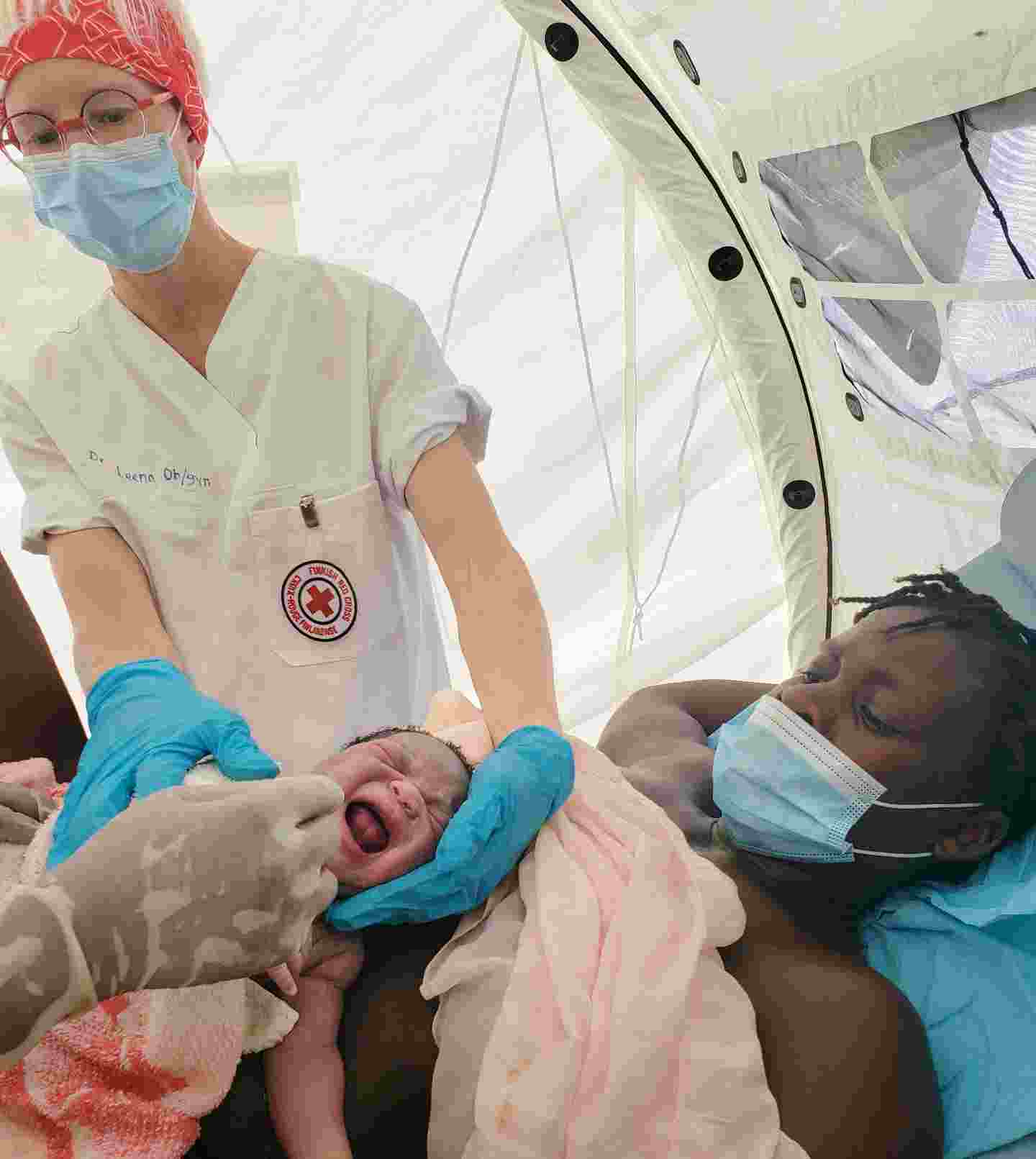 A doctor wearing Red Cross clothing examines a newborn baby held by its mother.