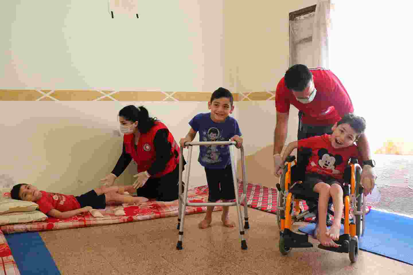 Three smiling Syrian children being helped by two Red Crescent employees.