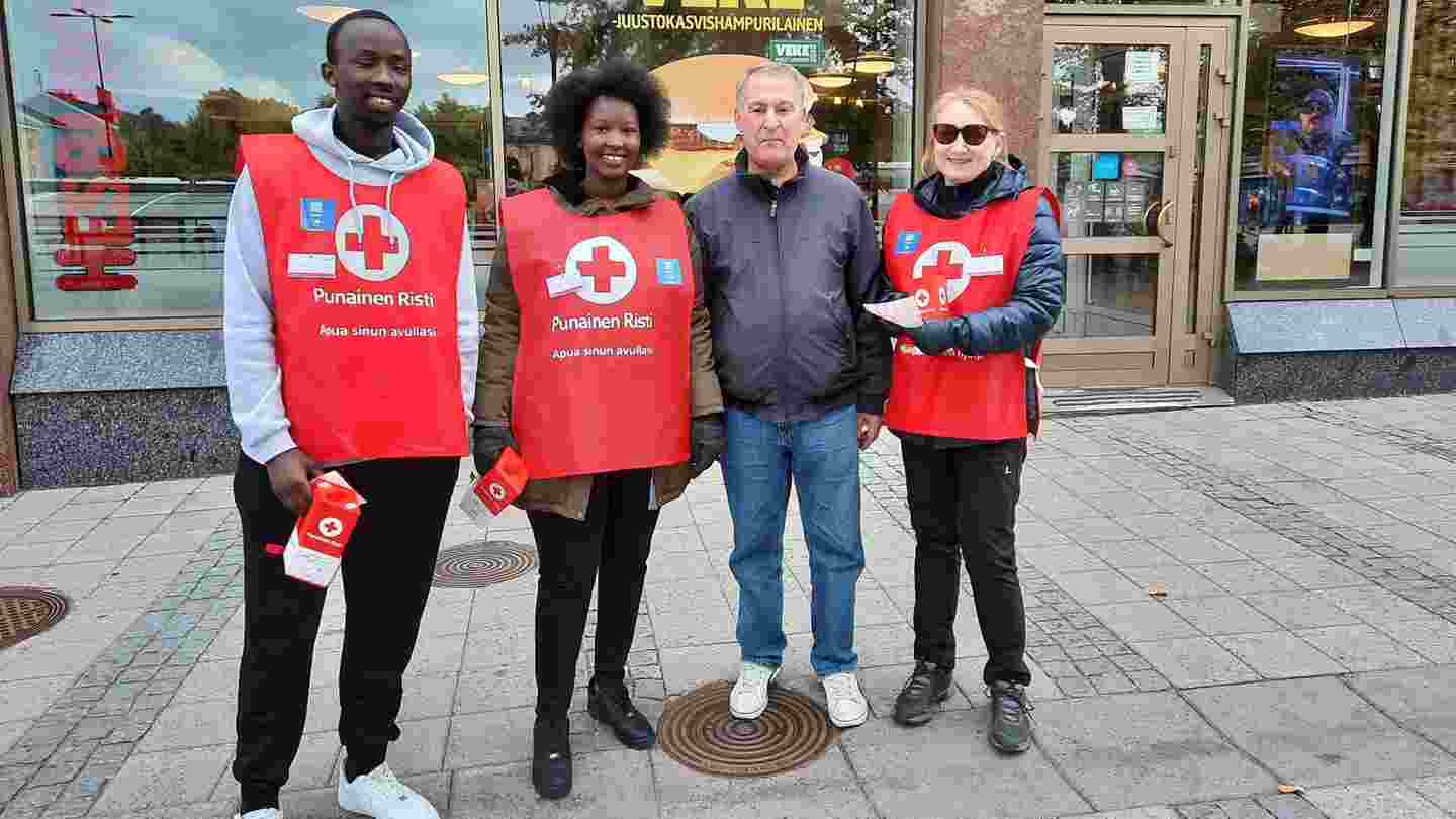 Four smiling people standing in front of a shopping centre. Two are holding a Hunger Day collection box, three are wearing red collecting vests of the Red Cross.
