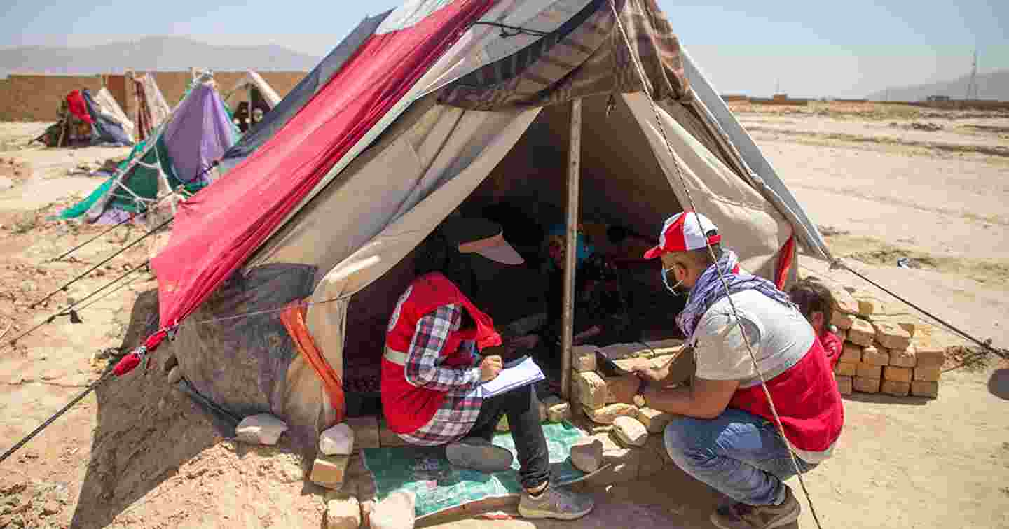Two Afghan Red Crescent employees talking to a person living in a tent.