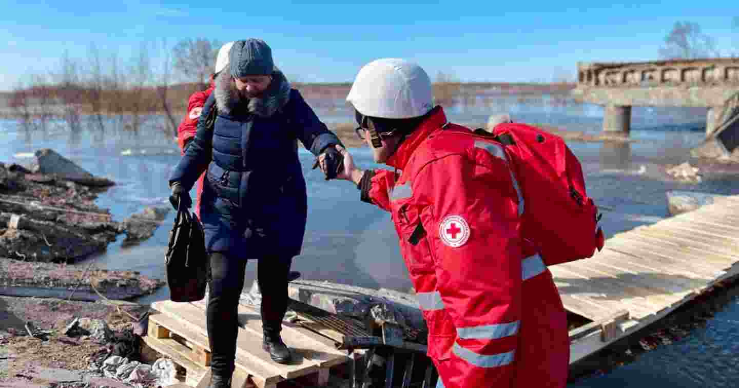 Red Cross volunteer helping an elderly person across a wooden bridge.