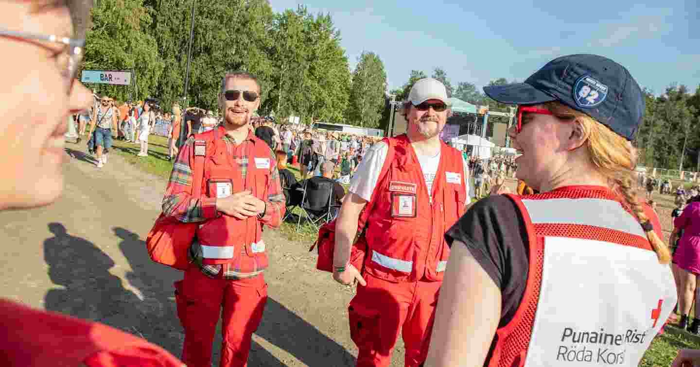 Four smiling Red Cross on-duty volunteers in a sunny festival area.