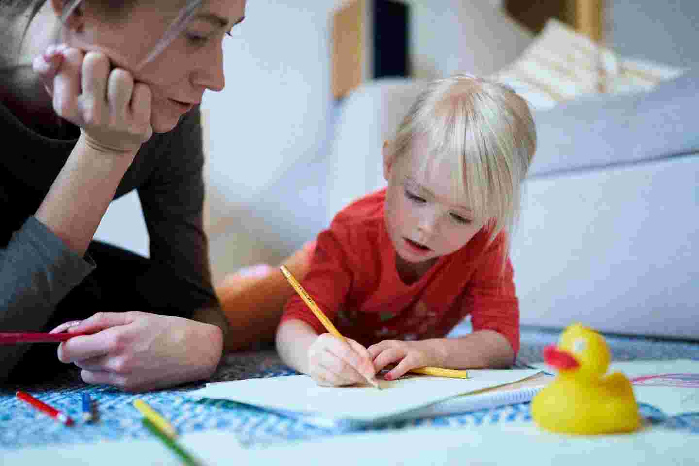 A child draws on a paper lying on the floor. An adult watches their drawing.