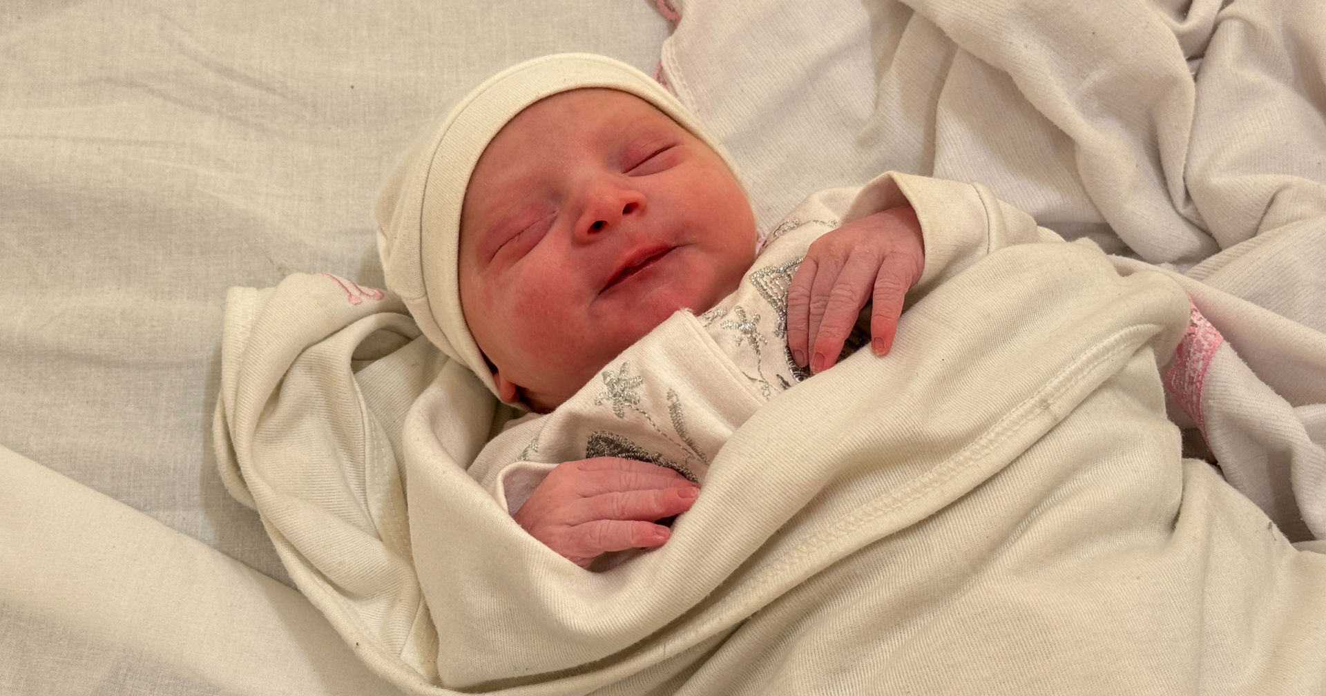 A newborn baby lies with eyes closed, wearing a white hat and a content expression on their face.