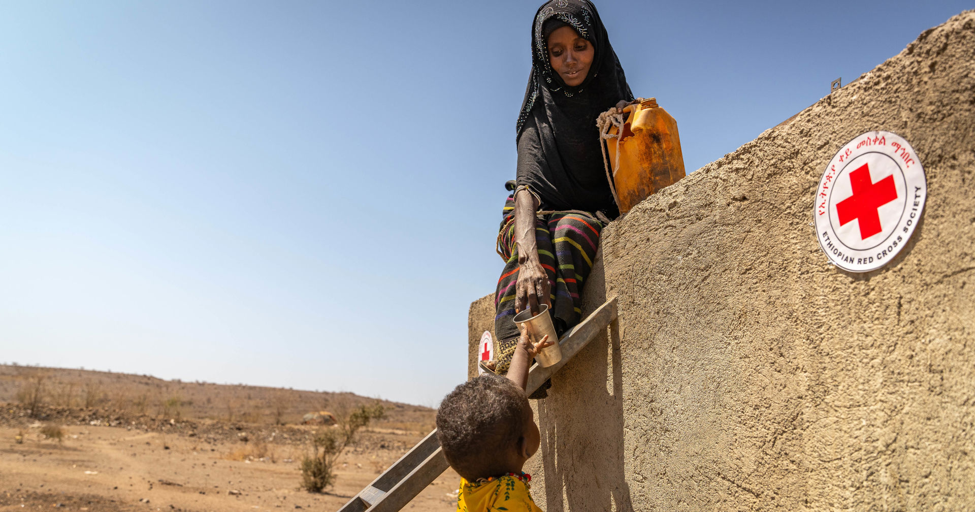 A woman wearing a scarf is handing water to a child waiting below.