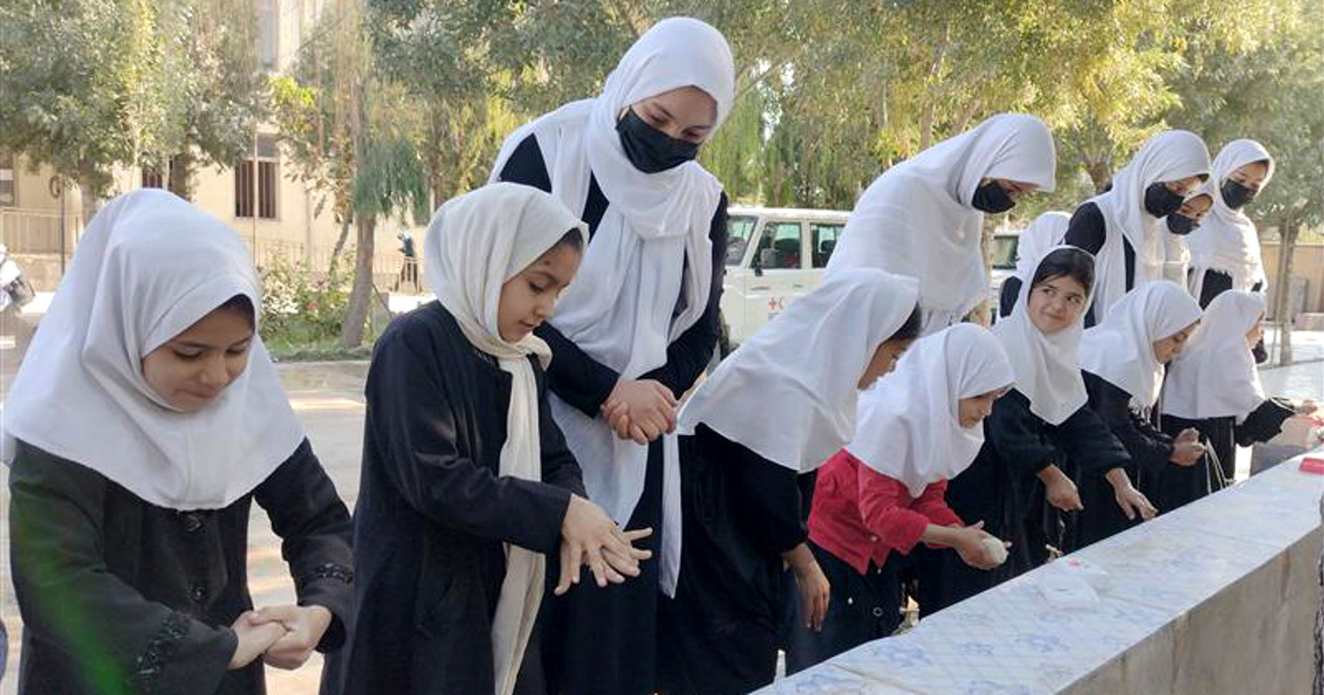 Girls are washing their hands in a row outside at a hand-washing station under the guidance of teachers.