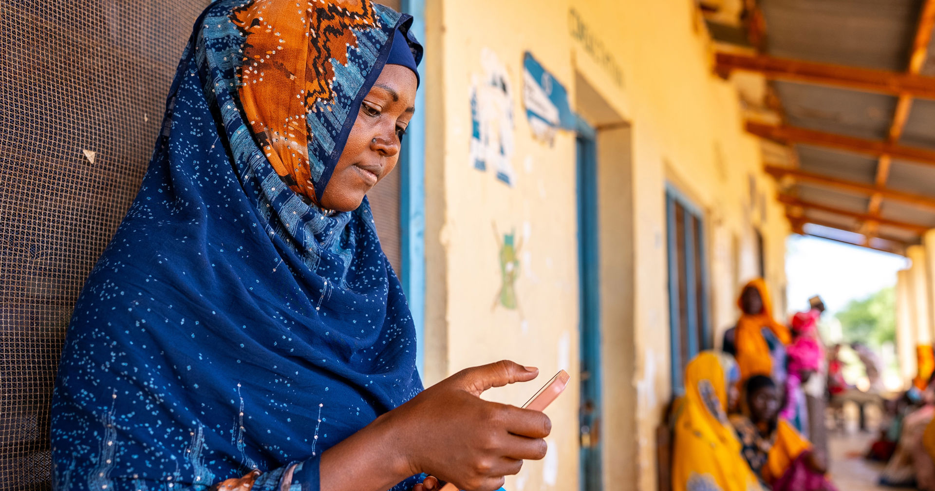 A volunteer wearing a blue scarf is looking at her mobile phone on the porch of a health clinic.