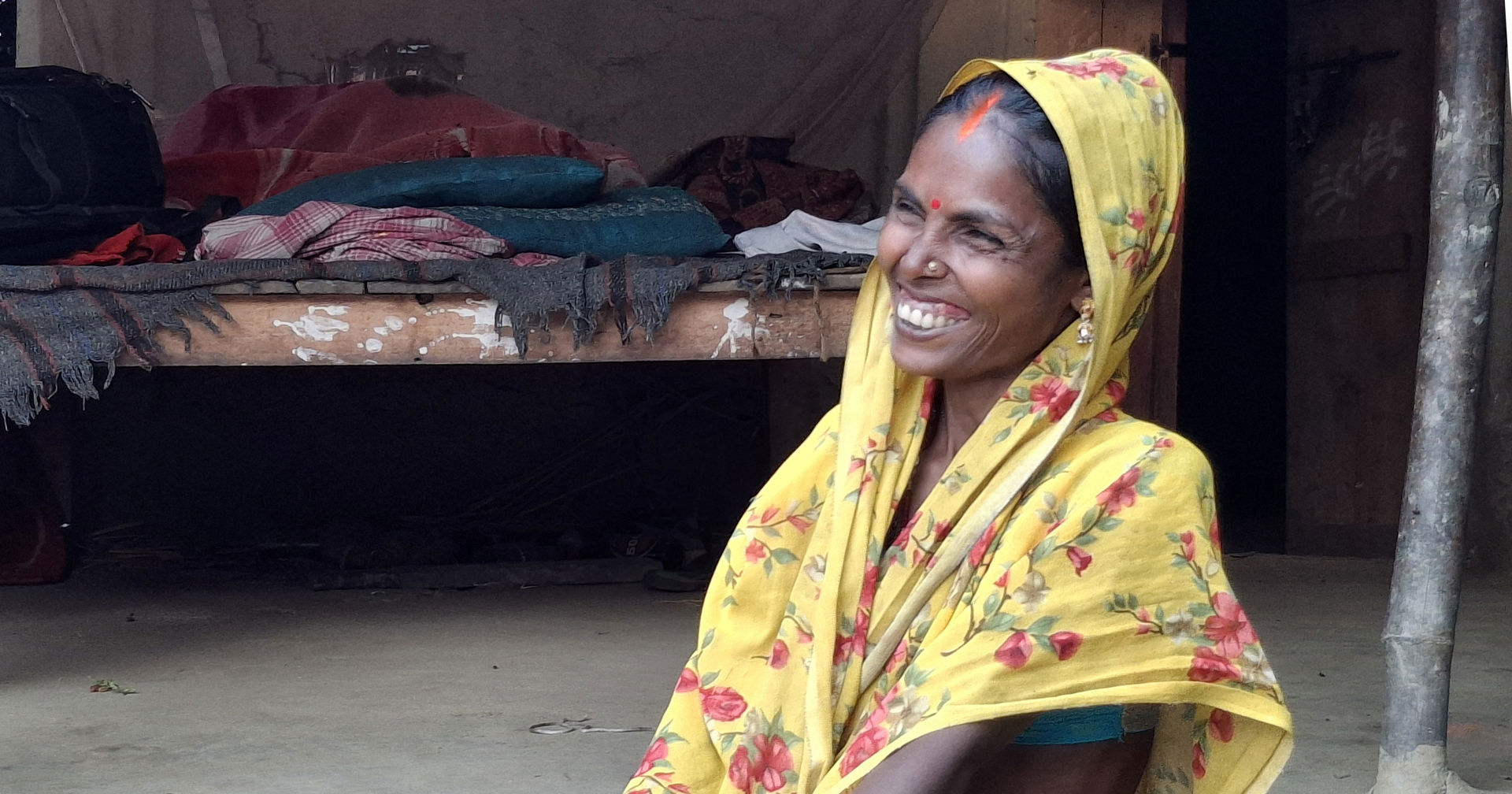A woman wearing a yellow sari is smiling while sitting on the porch of her home.