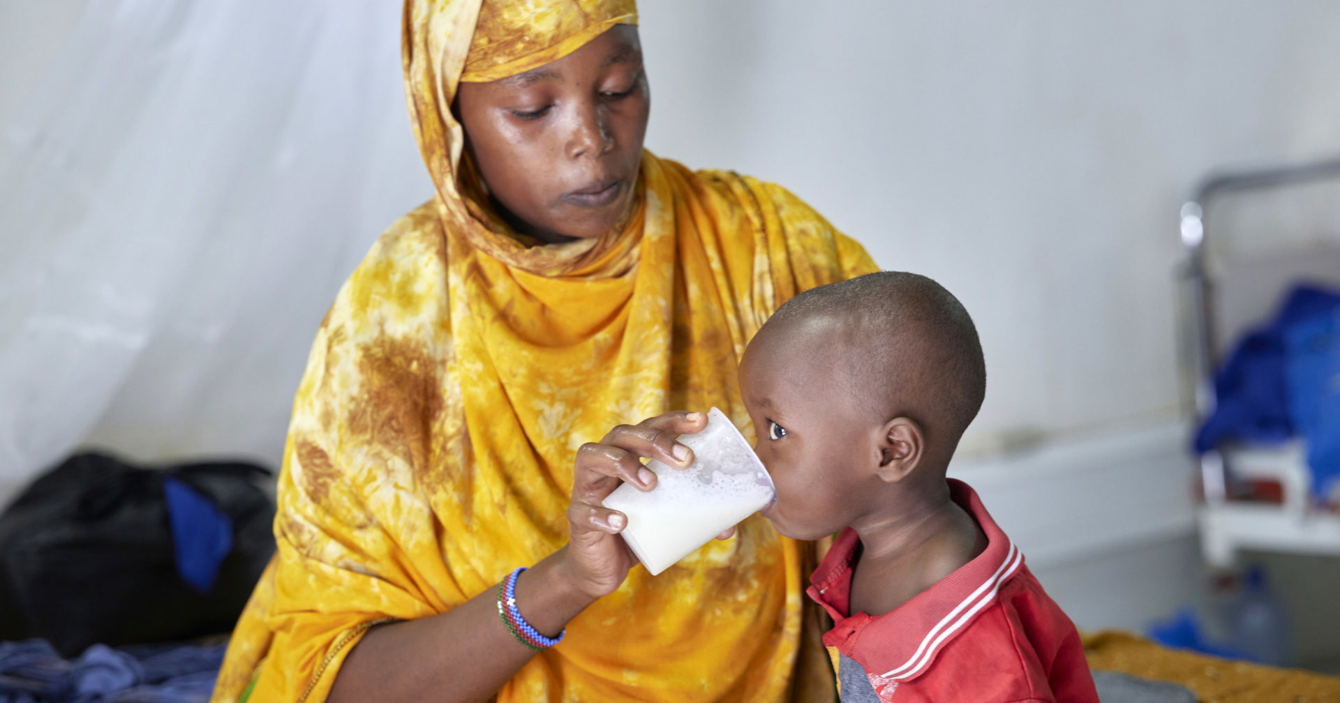 A mother is sitting on a hospital bed and feeding milk to a child.