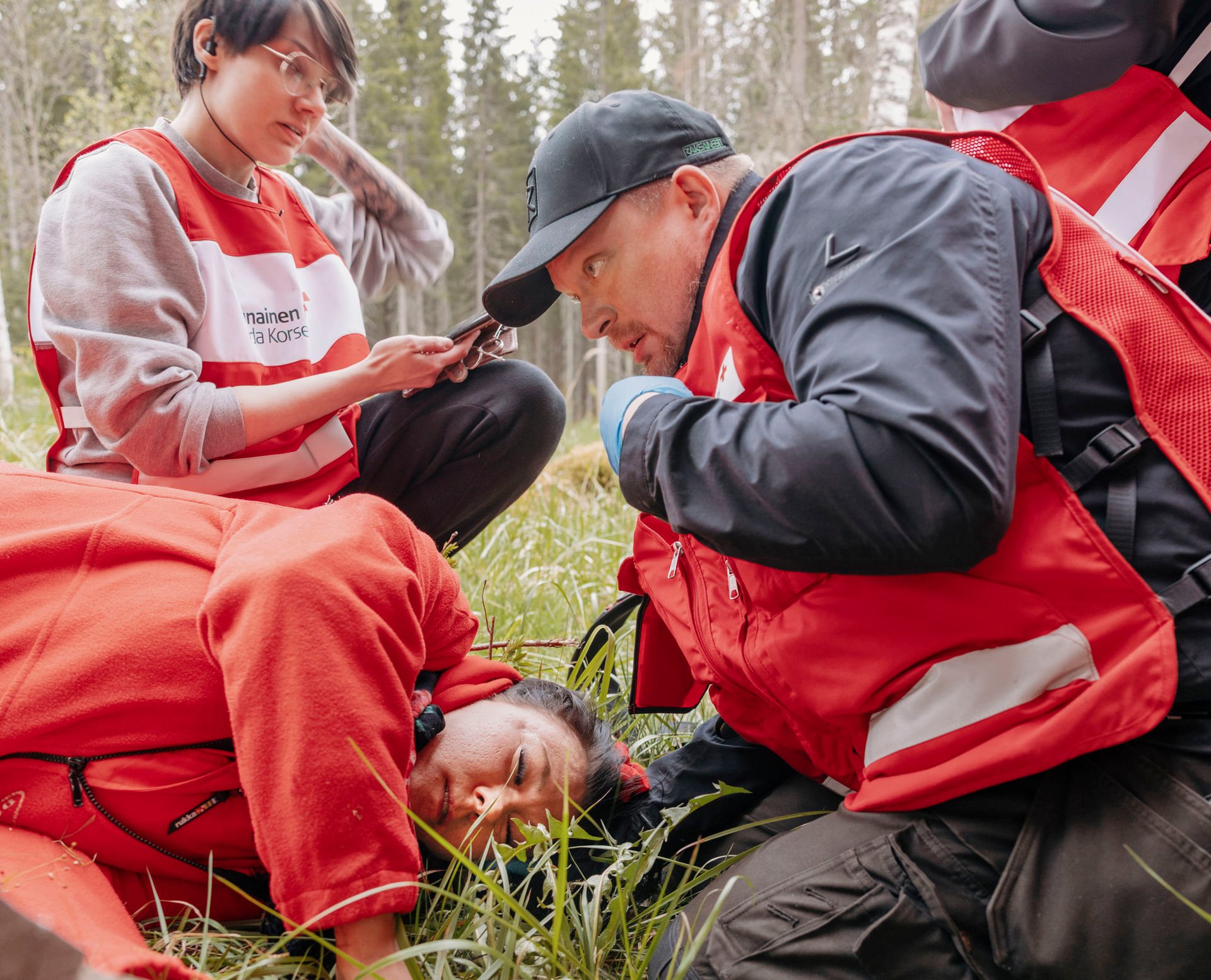 Red Cross volunteers practise first aid situations in wooded terrain. One person lies on the ground in a red coat, simulating an injured person, while another volunteer checks the situation and a third uses a telephone. In the background you can see trees and green grass.