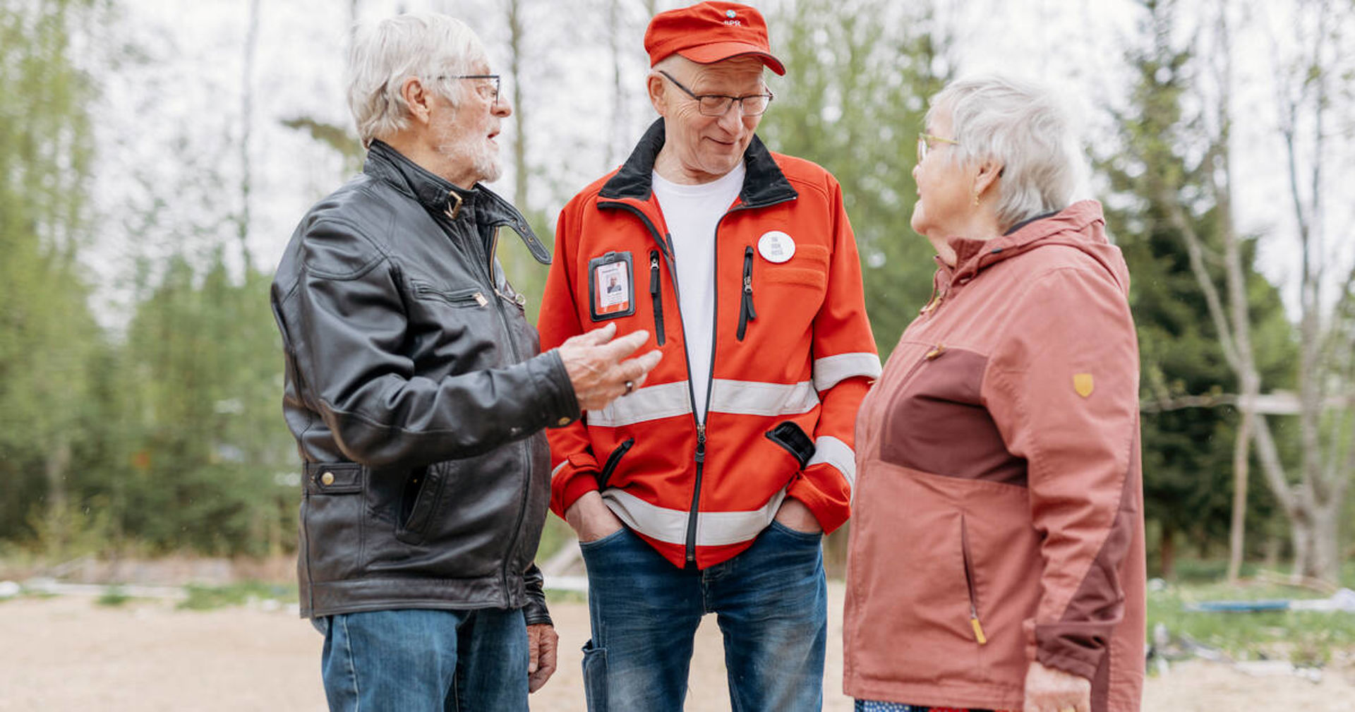 Three people are standing outside talking. The person in the middle is wearing a red Red Cross vest and cap, with an identification card on his chest. In the background you can see green trees and sandy soil.
