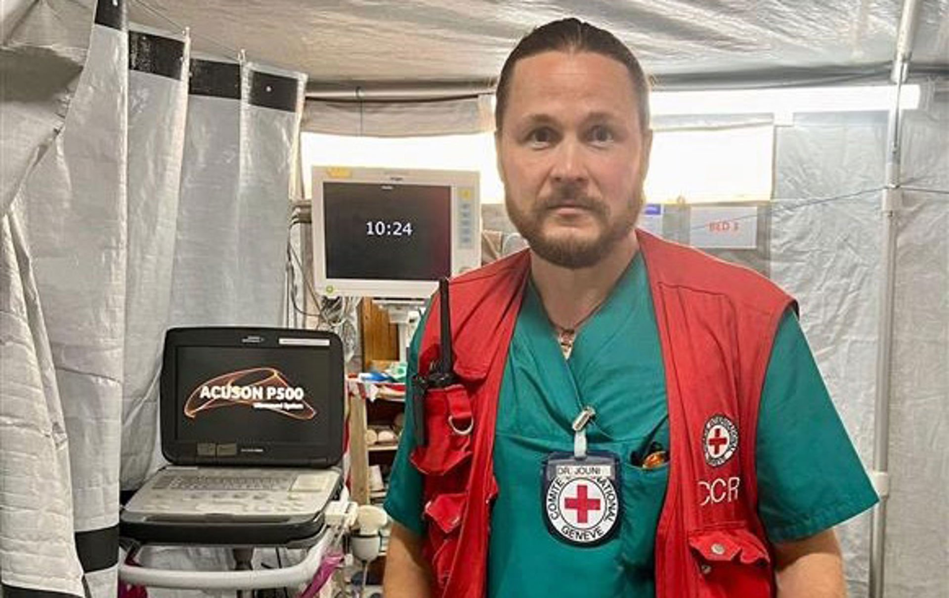 A person is standing in a field hospital tent with hospital equipment. He is wearing a green hospital gown and a Red Cross vest with the organisation's emblem and an International Committee of the Red Cross identification card. In his breast pocket he carries a walkie-talkie.