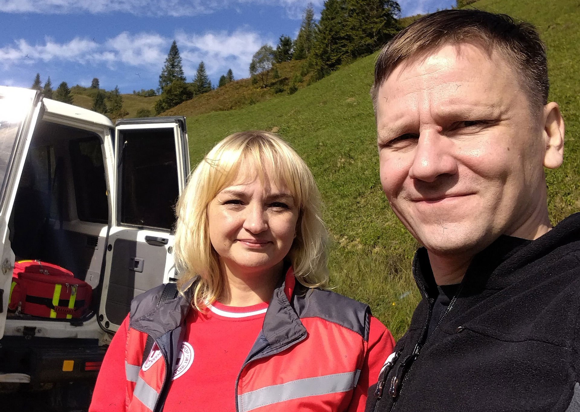 Two people are standing outside on a green slope. One of the people is wearing Ukrainian Red Cross work clothes. In the background you can see a pine forest and a blue sky. A white van with its rear doors open is parked next to the people, and inside you can see a large red bag of medical supplies.