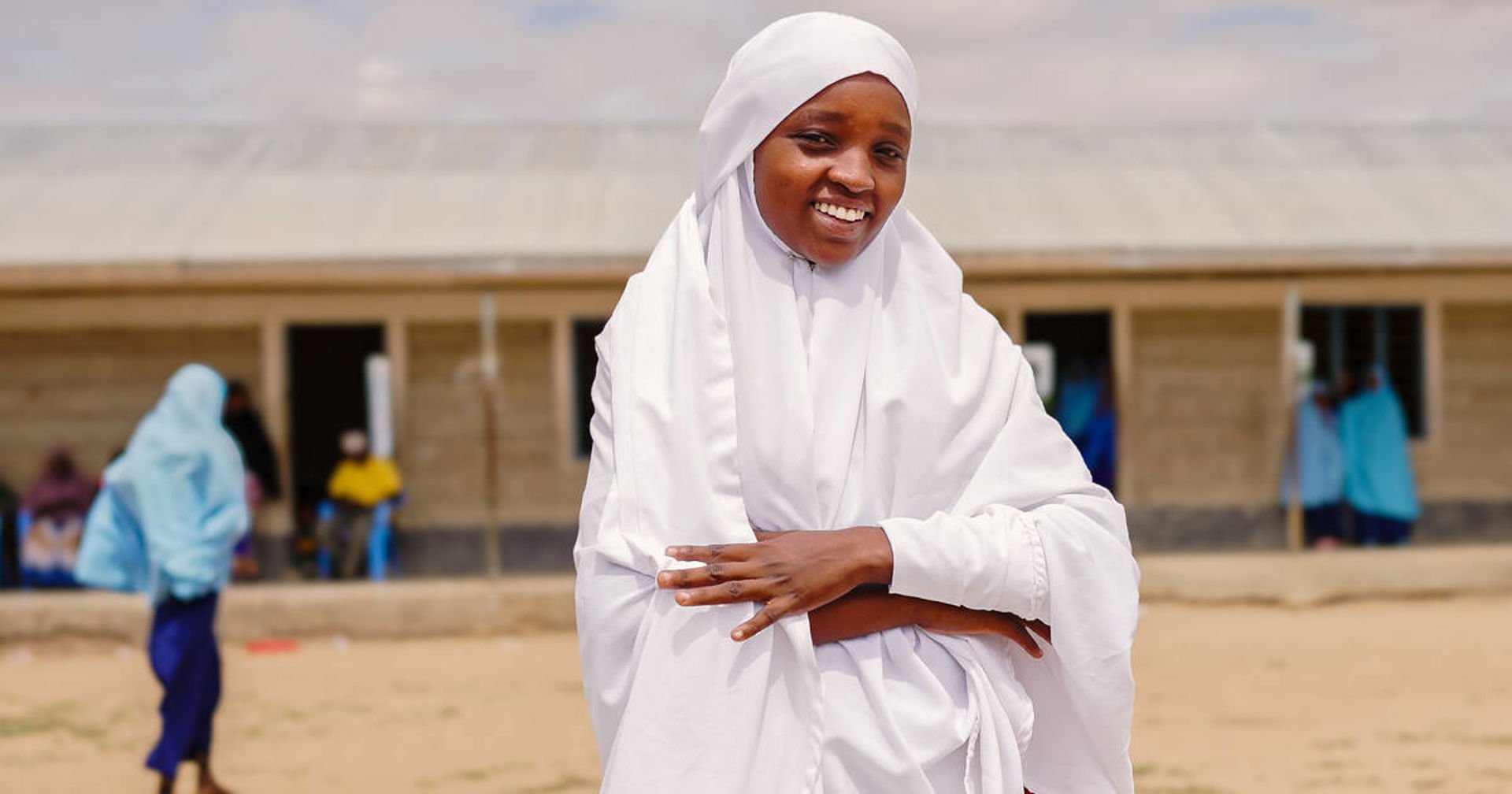 A person dressed in white is standing outside in a sandy area, with a single-storey school and pupils in blue school uniforms in the background.