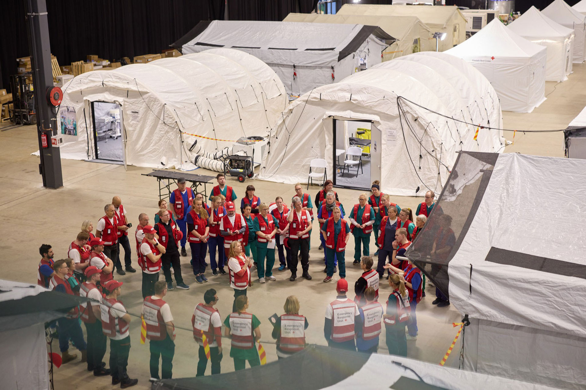 A large indoor space with several white tents set up in rows. In the foreground, a large group of people stand in a circle, dressed in Red Cross work vests. More tents, boxes and equipment, such as a generator, can be seen in the background. The floor is concrete and the space is well lit. This is a field hospital set up in an exhibition centre, where Red Cross staff meet to receive instructions.