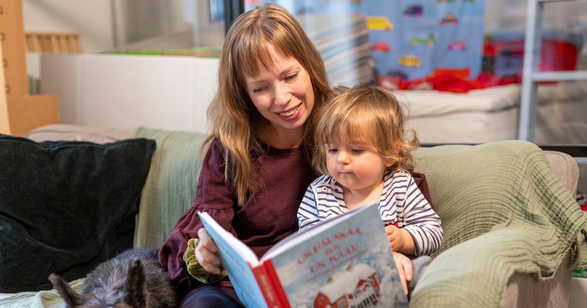 A smiling adult sits on a sofa reading a book to a small child on her lap. The book is called ‘It's fun when it's Christmas’ and has a colourful cover with a winter landscape with red buildings. A small dog is lying on the sofa. Toys and colourful pictures on the wall in the background create a cosy atmosphere.