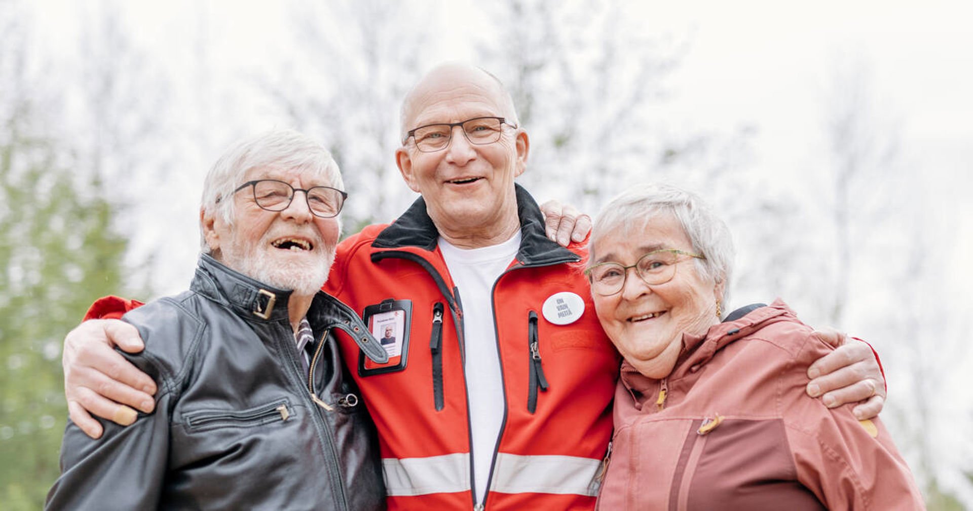 Three smiling people standing outside. The person in the middle has his hands on the shoulders of those at his sides. The person in the middle is wearing a red Red Cross vest with the organisation's emblem. In the background you can see a pale sky and deciduous trees.