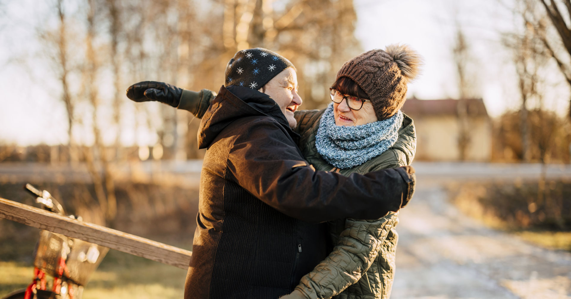 Two smiling people hugging outside in a winter landscape. Both are wearing winter clothes, such as thick coats, hats and scarves. Bare trees and a light-coloured building can be seen in the background, and there is a thin layer of snow on the ground.