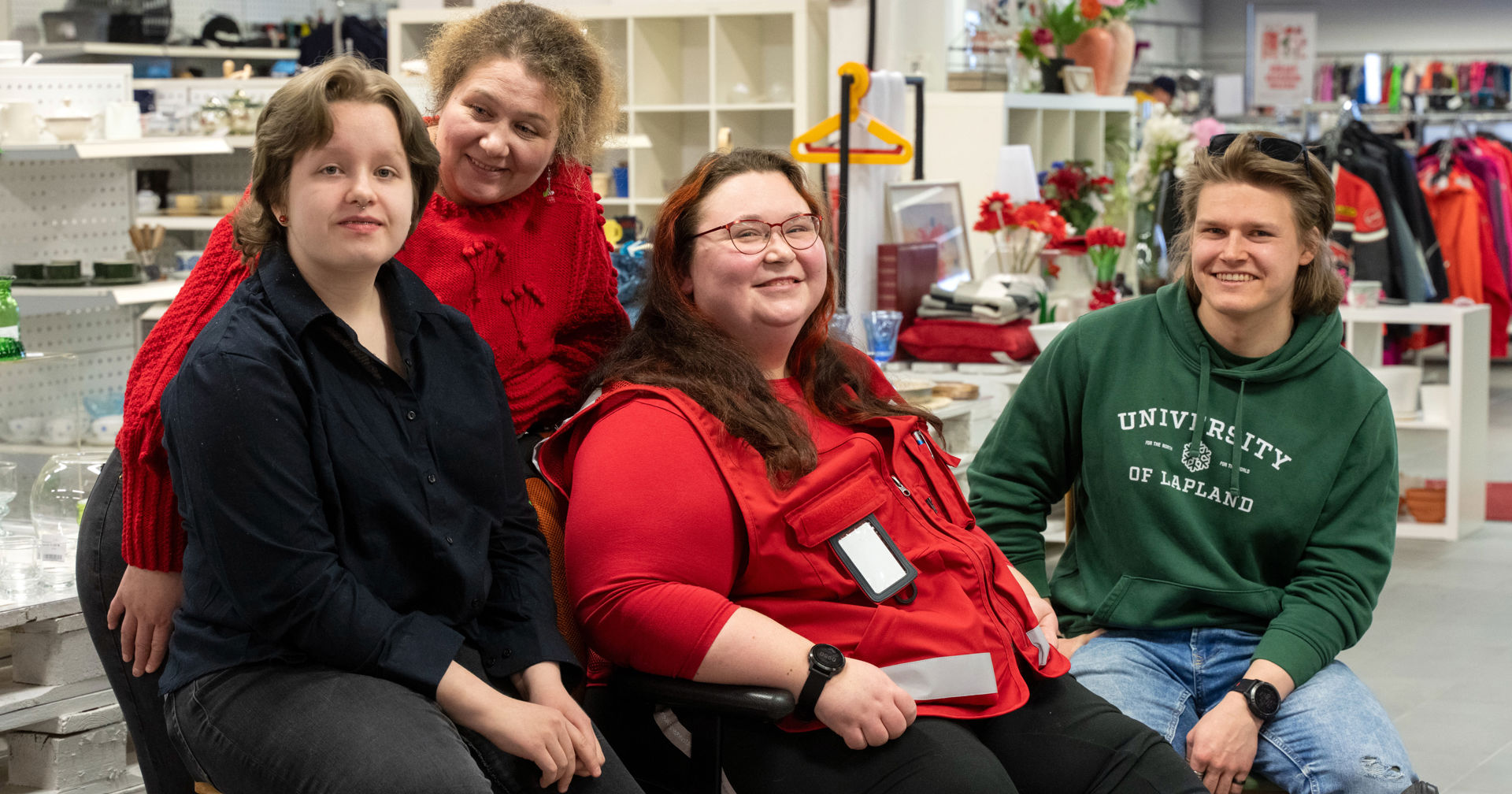 A smiling Red Cross volunteer sits inside, wearing a red vest with an identification card. In the background you can see shelves, red decorative flowers, clothes and household items, suggesting a flea market or donation space.