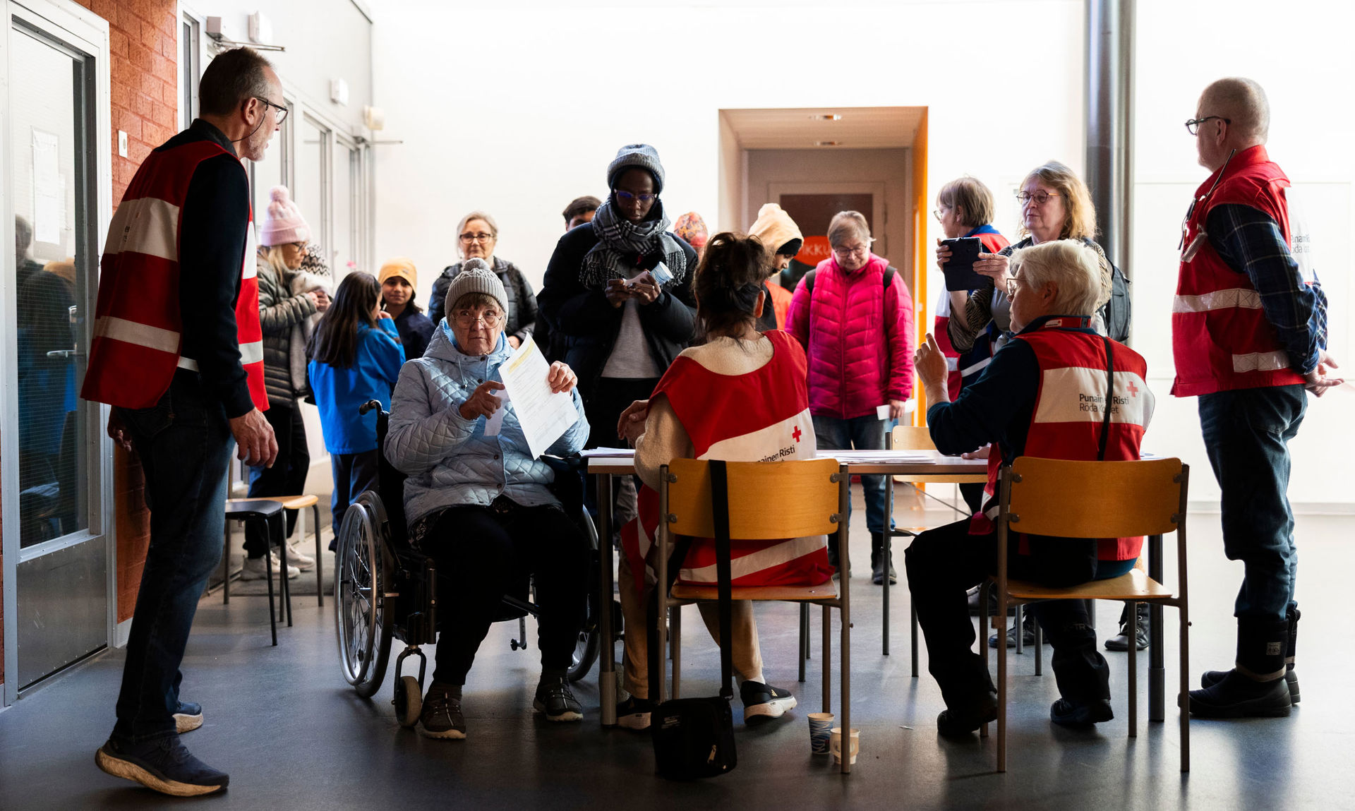 An indoor situation where volunteers and people gather around a table. At the front, two people sit on chairs and one in a wheelchair, with papers and pens on the table. They are wearing Red Cross volunteer vests. In the background, several people stand in a queue or group, dressed in outdoor clothing such as jackets and beanies. The space has a light wall and a glass door on the left, with lighting from above and outside.