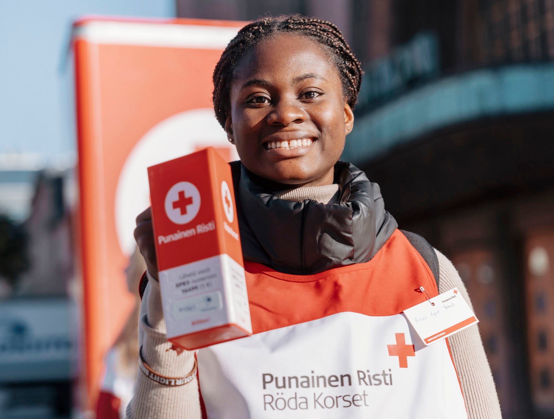 A smiling volunteer holds a collection box for the Hunger Day collection. The person is wearing a Red Cross volunteer vest with the Red Cross logo and the text ‘Red Cross Röda Korset.’ In the background, you can see a large Red Cross collection box and the surrounding cityscape.