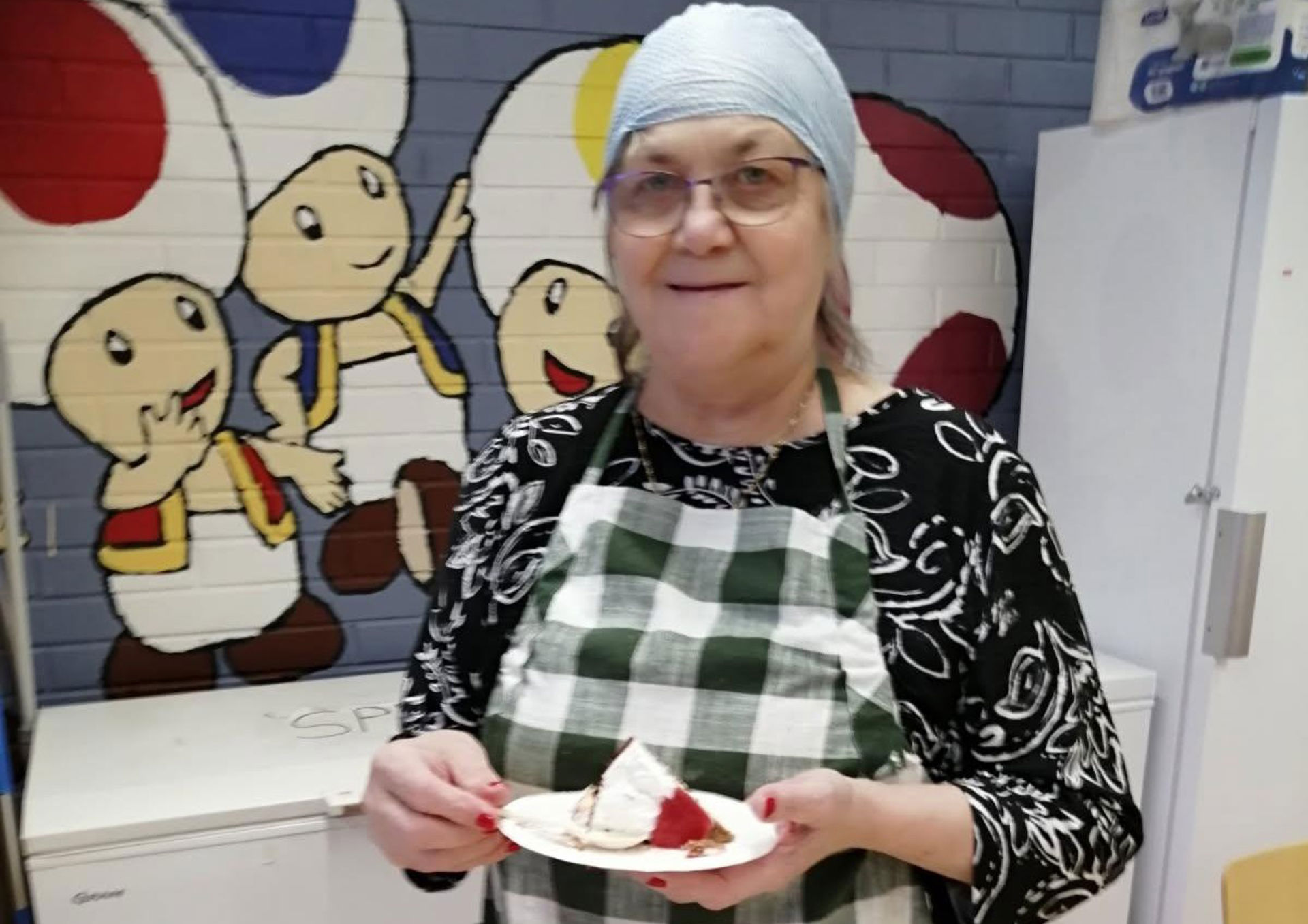 A Red Cross volunteer stands indoors during a food aid distribution event, wearing a green and white checkered apron and a hair restraint. She is holding a plate with a piece of cake and whipped cream. In the background you can see a colourful mural with cartoon characters, white cabinets and a fridge.