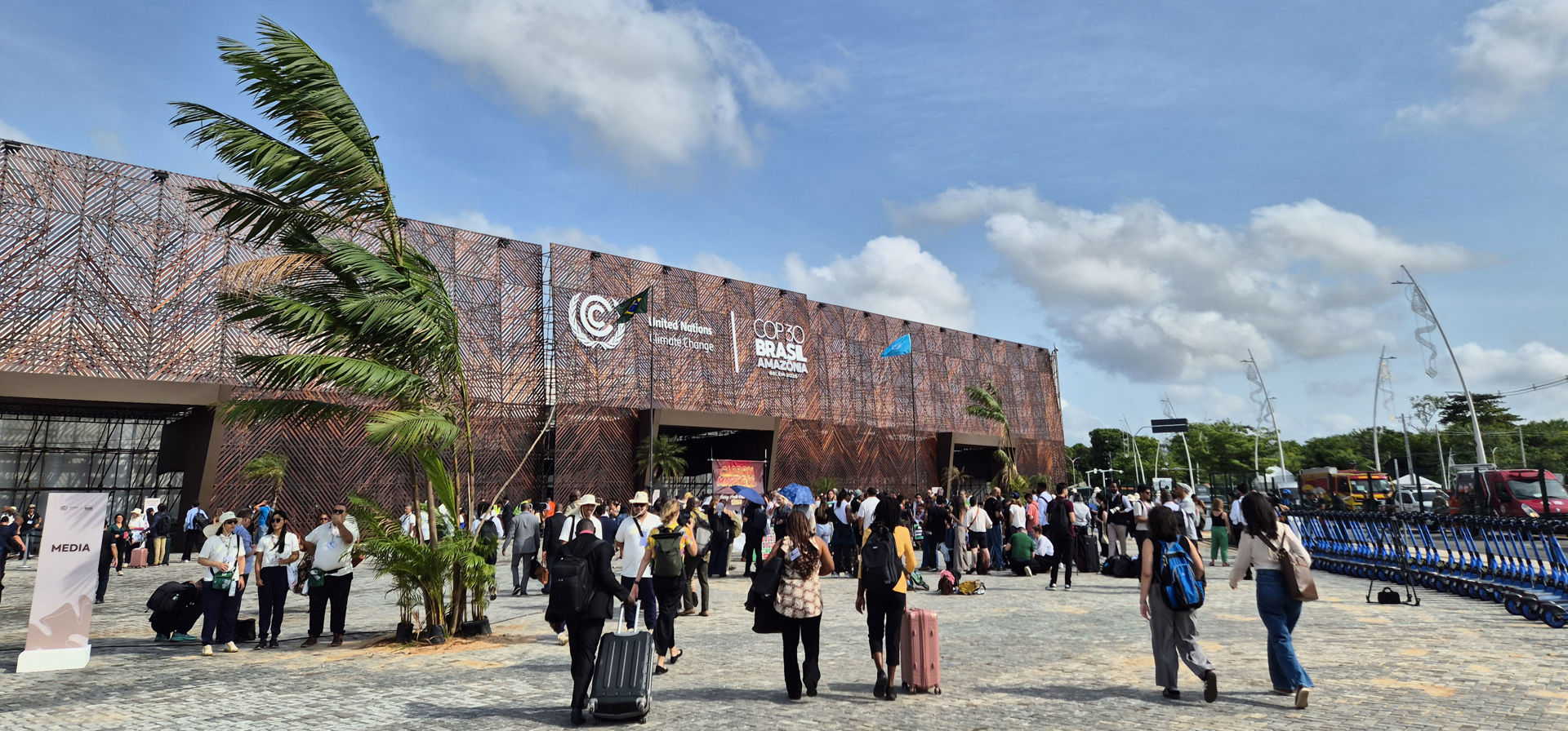 People gathered in front of a large event building on a sunny day. The building’s facade displays the UN Climate Change Conference logos and the text ‘COP30 Brazil Amazonia Belém 2025.’