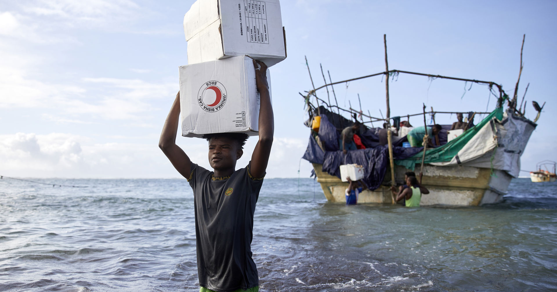 A Red Cross volunteer wading in the sea, carrying Red Cross aid boxes on his head.