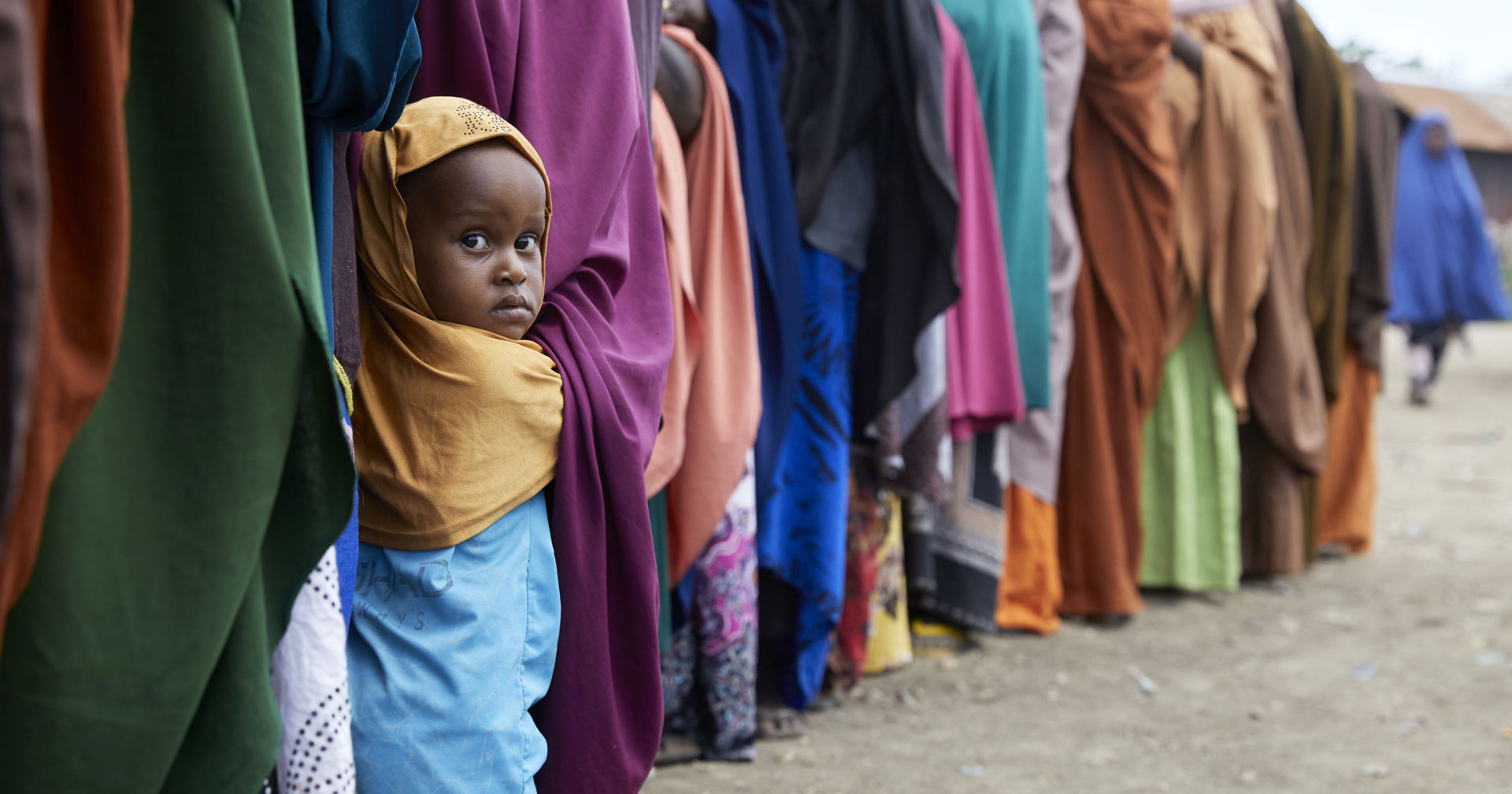 A child looks at the camera in a food queue with lots of adults queuing.