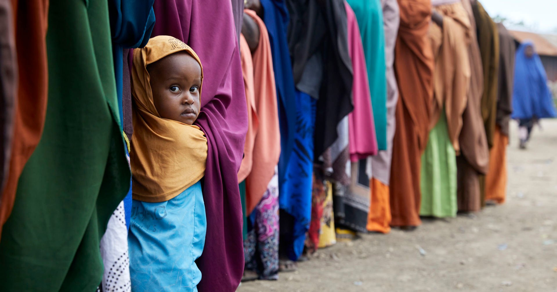 A child looks at the camera in a food queue with lots of adults queuing.