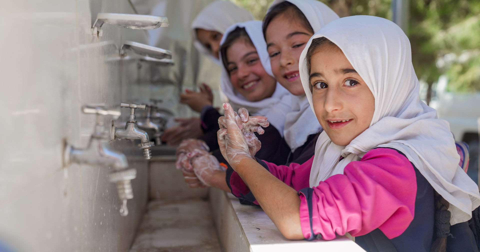 A group of pupils wearing white scarves are standing in a line, lathering their hands with soap at an outdoor hand-washing station. 