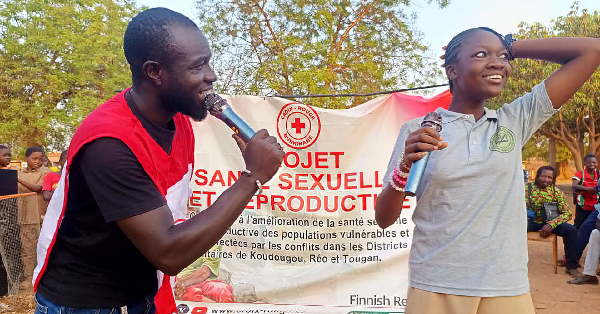 Two people holding microphones are standing outdoors in front of a Red Cross poster. One of them is wearing a Red Cross vest and speaking into their microphone. 