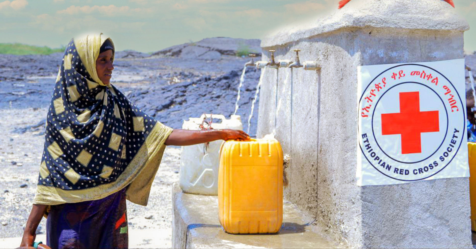 A person wearing a blue and yellow scarf is standing outside at a water station, pouring water into a yellow canister next to the Ethiopian Red Cross emblem. 