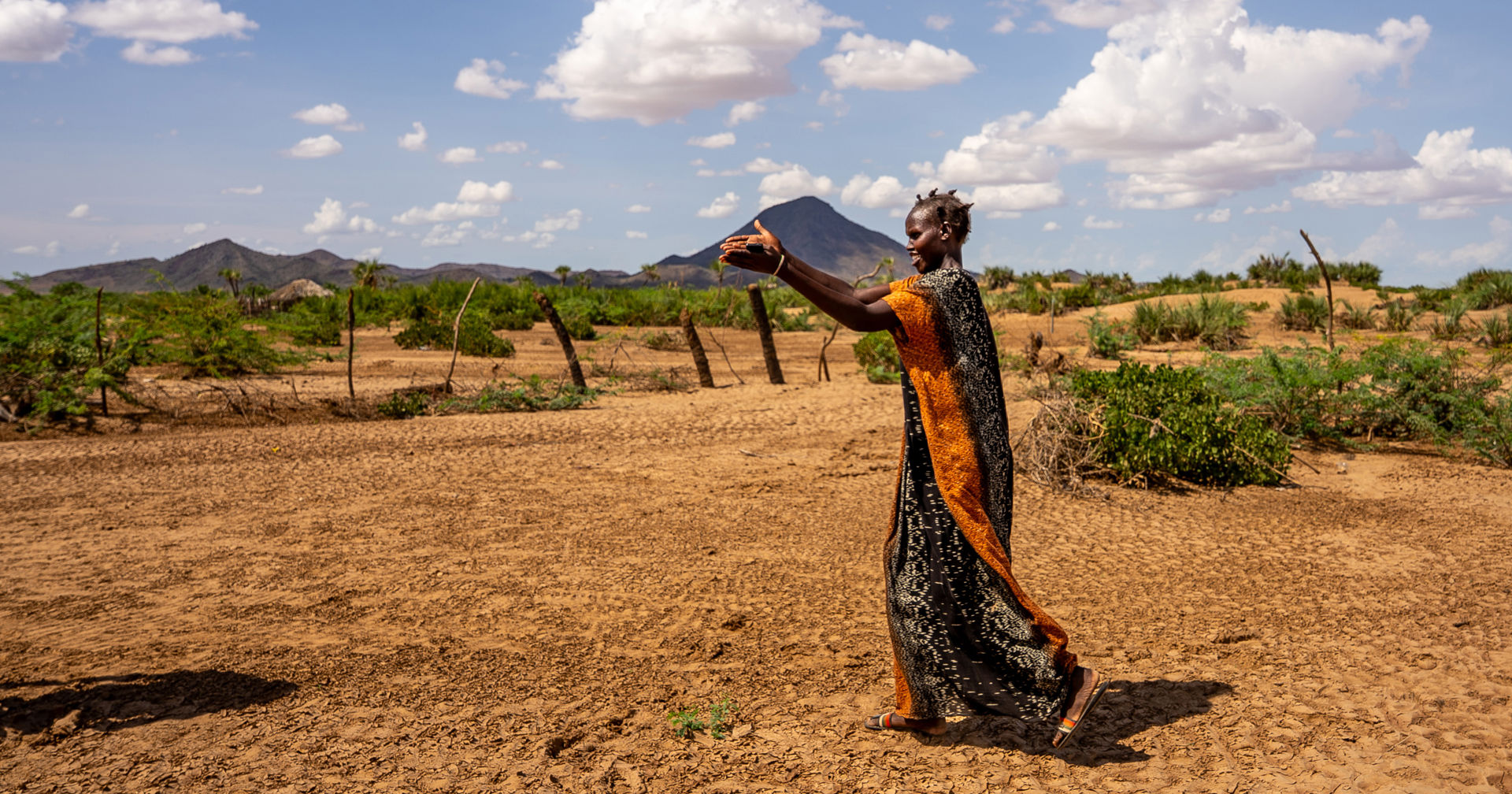 A person standing on brown sand outside in the sunshine. They have lifted their hand up and are pointing towards something in the distance. 