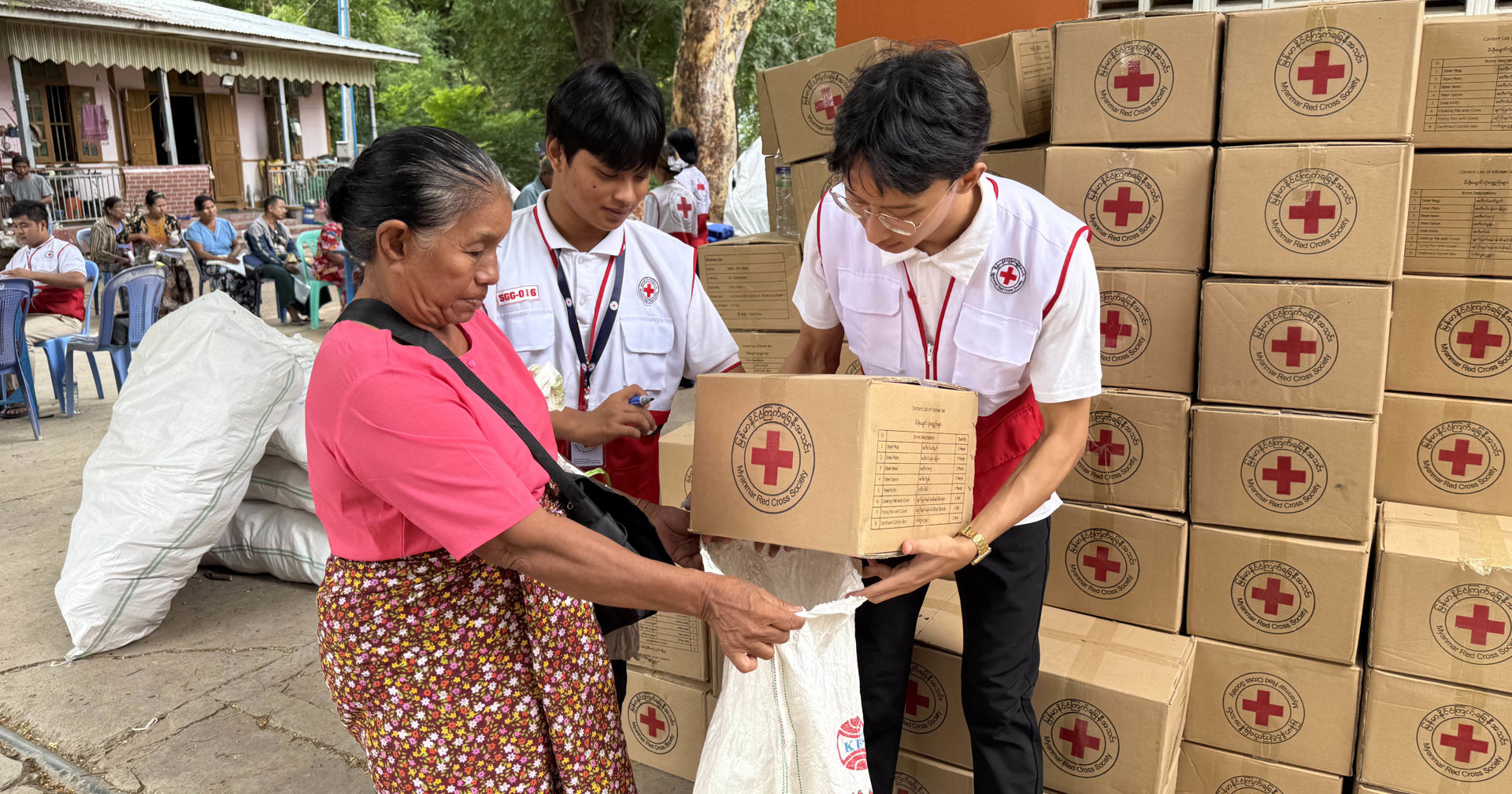 Two people dressed in Red Cross clothes are placing a cardboard box bearing the Red Cross symbol into a plastic bag being held by an elderly person. 