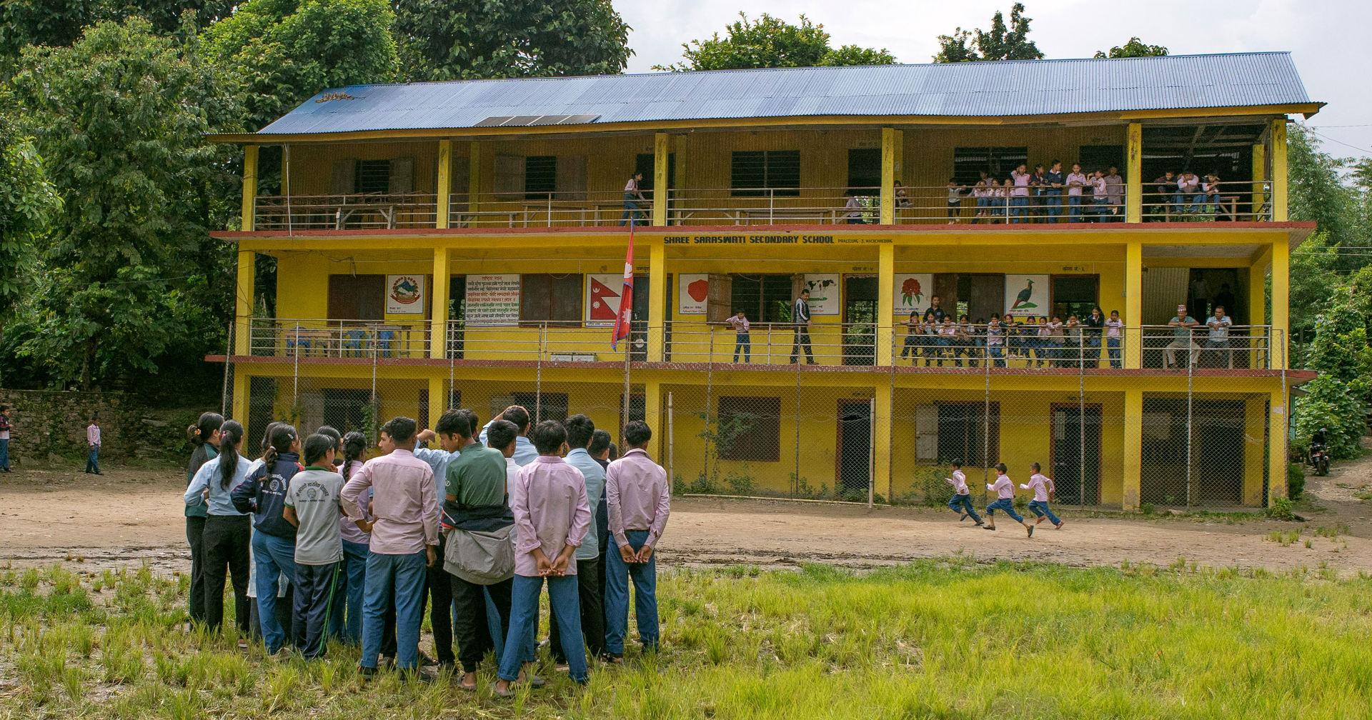 A group of students are standing in the yard of a yellow school building. Some of the children are running across the yard.