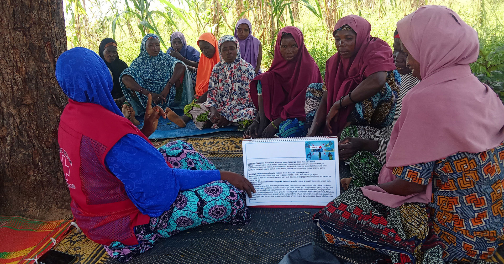 A group of people wearing scarves are sitting in a circle outdoors on mats, looking at a paper with some text on it in the middle. The person holding the paper is wearing a Red Cross vest. 