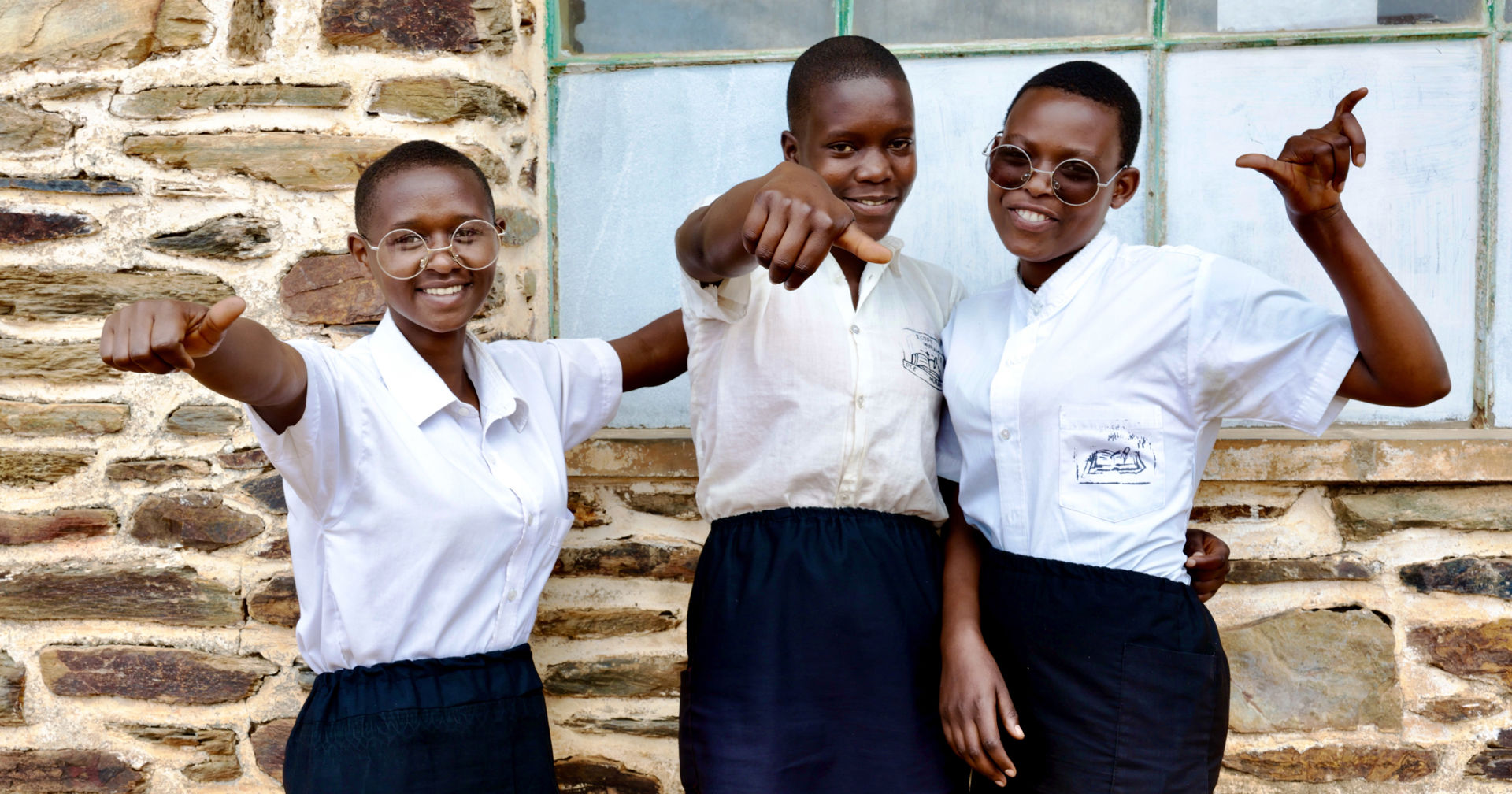 Three students wearing white collared shirts and black skirts are cheering outside. There is a stone wall and a window behind them. 