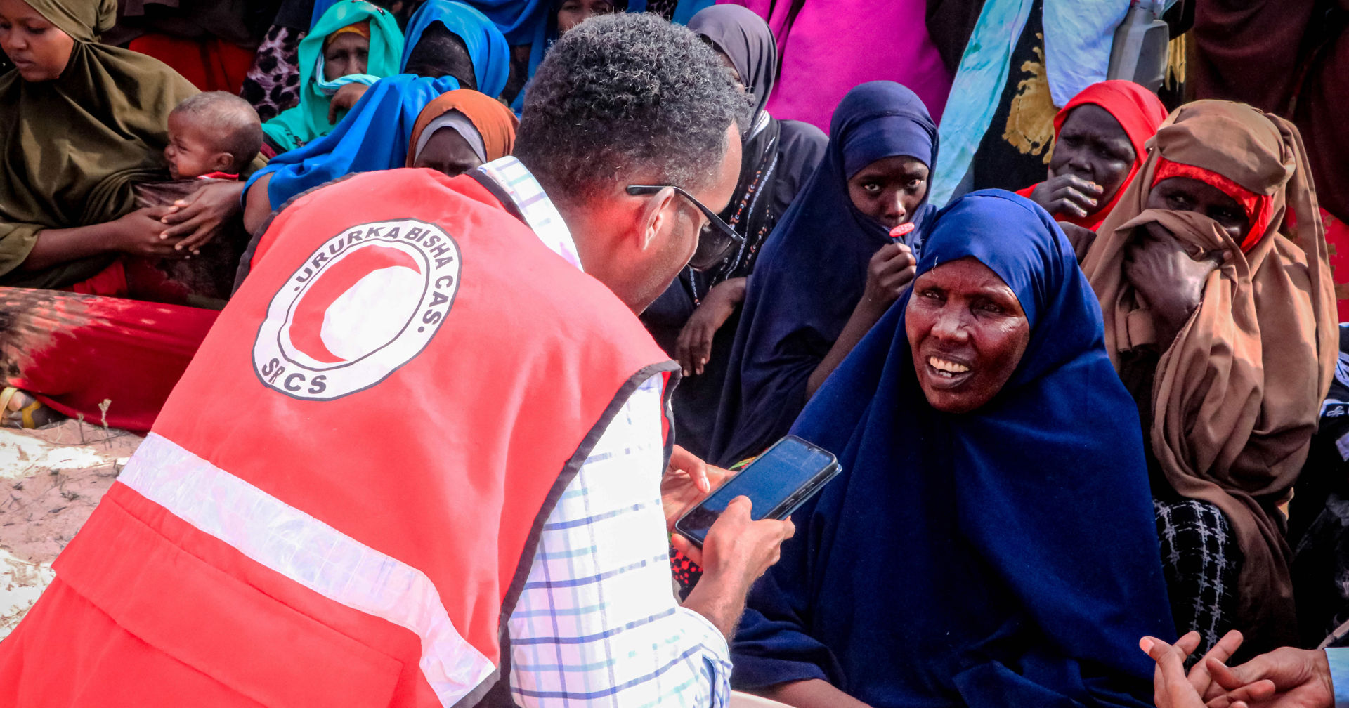 A person wearing a Red Crescent vest is crouching outside in the sunshine in front of a person wearing a scarf and tapping on their mobile phone. 